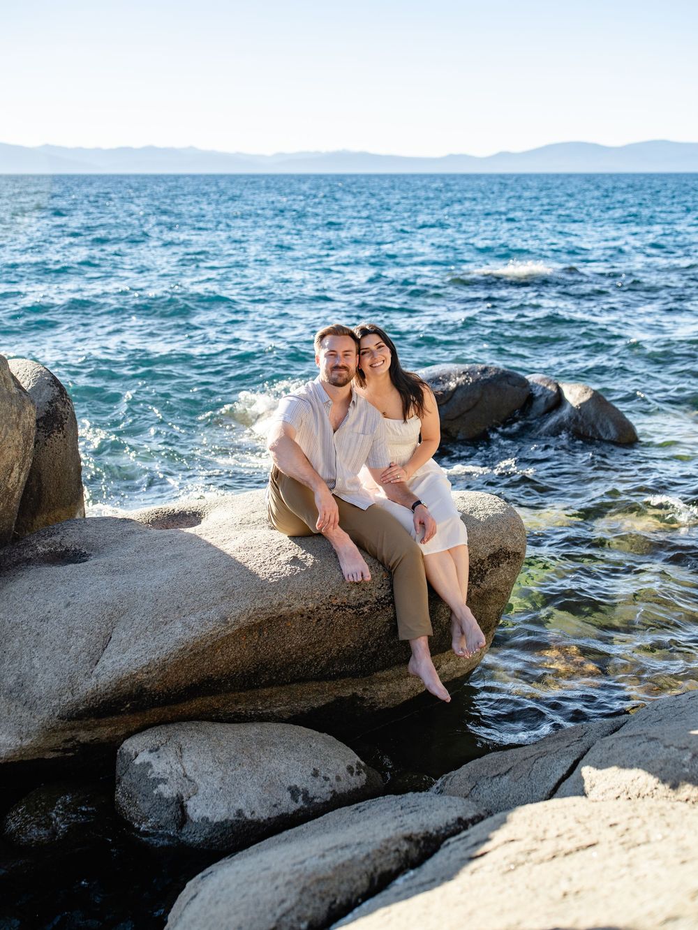 Couple relaxes on large granite boulders by the blue waters of Lake Tahoe during a sunny afternoon.