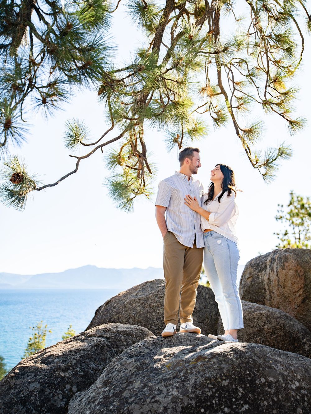 Couple poses together on rocky cliffs overlooking Lake Tahoe with pine trees framing the romantic scene.
