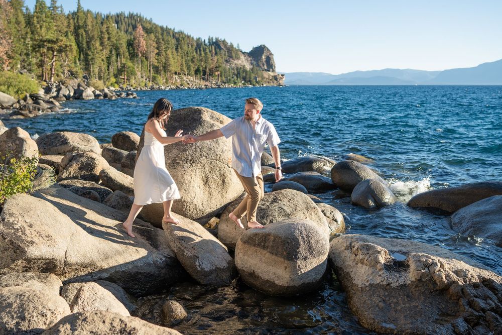 Panoramic view of couple sitting on rocky shoreline with mountains and forest surrounding Lake Tahoe.