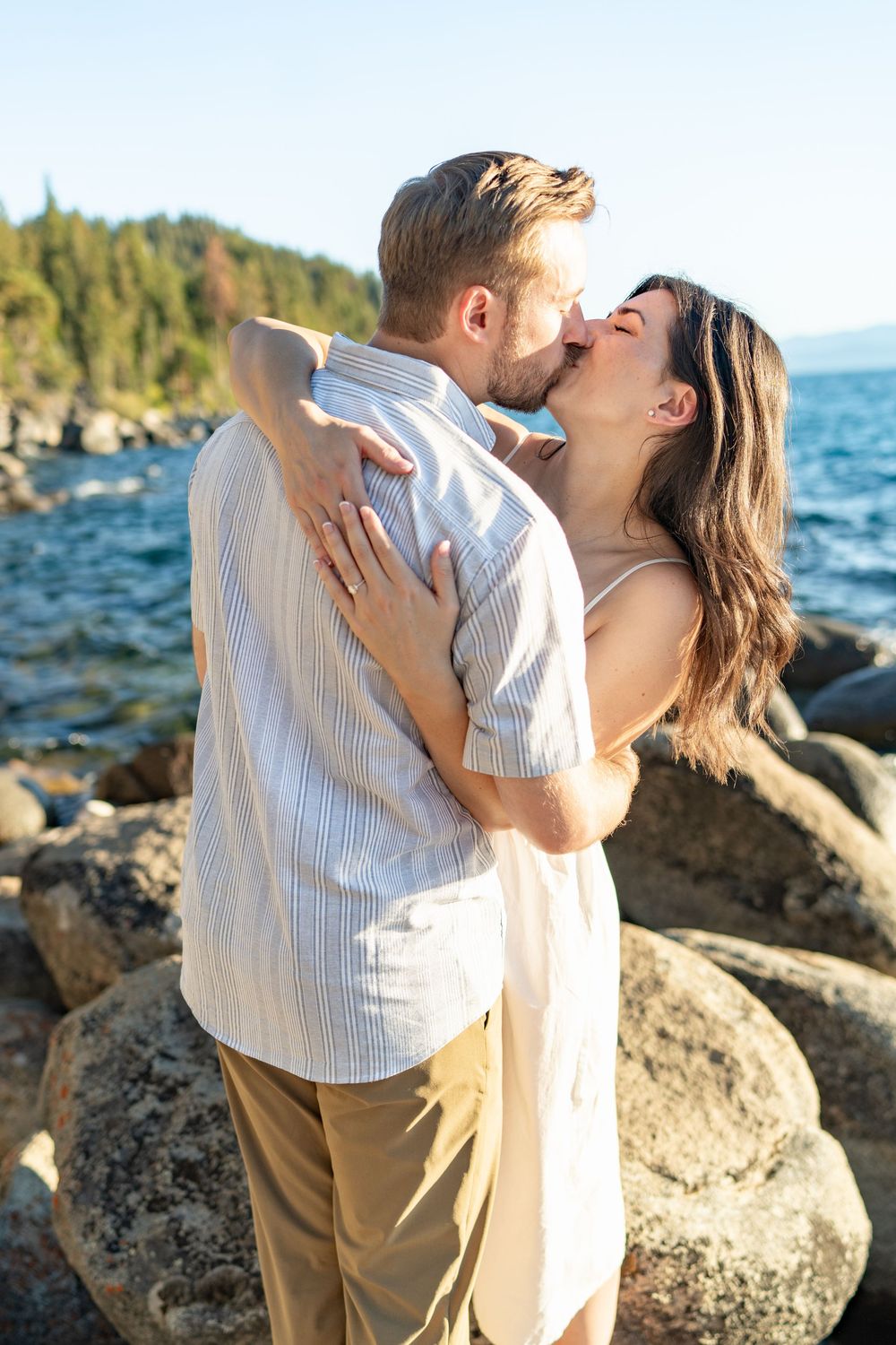 Romantic couple embraces on rocky beach at sunset with ocean and mountains in background.