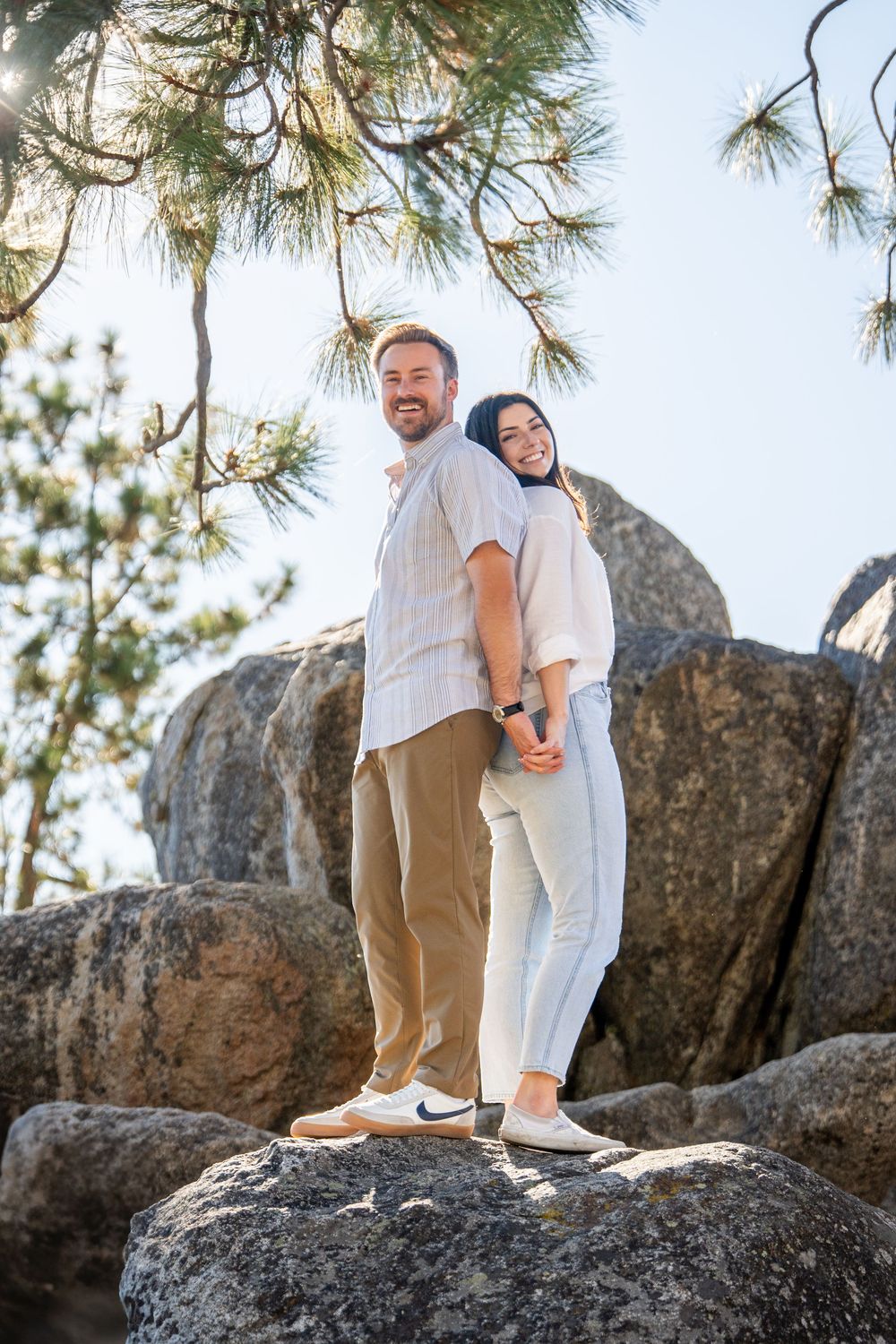 A couple stands together on rugged rocks with pine trees in the background during an outdoor photo session.