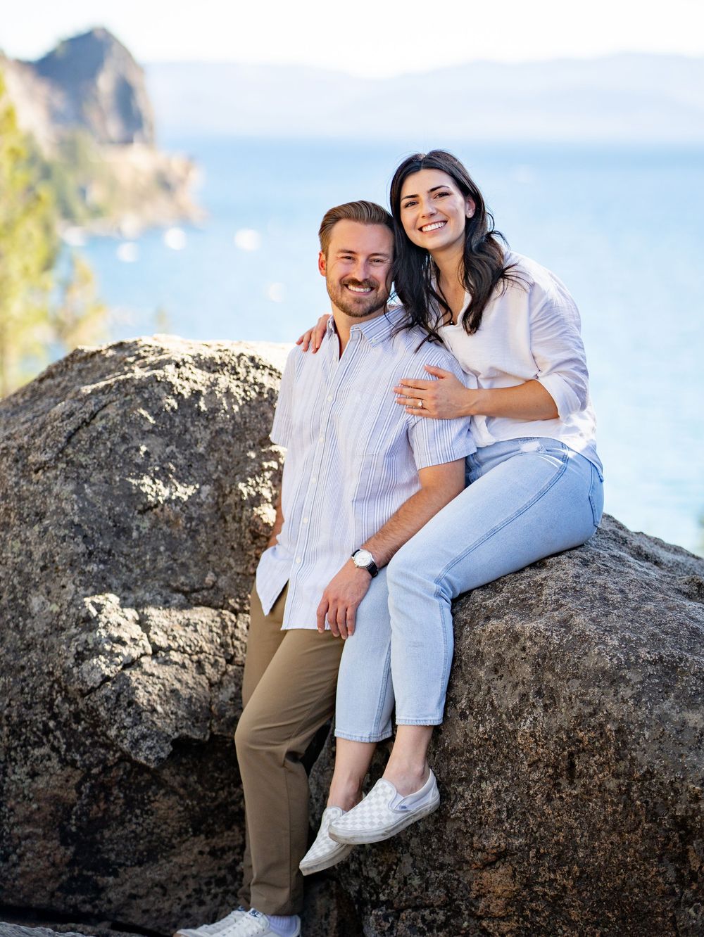 A couple shares joyful moments while sitting on large coastal rocks during a sunny beach photoshoot.