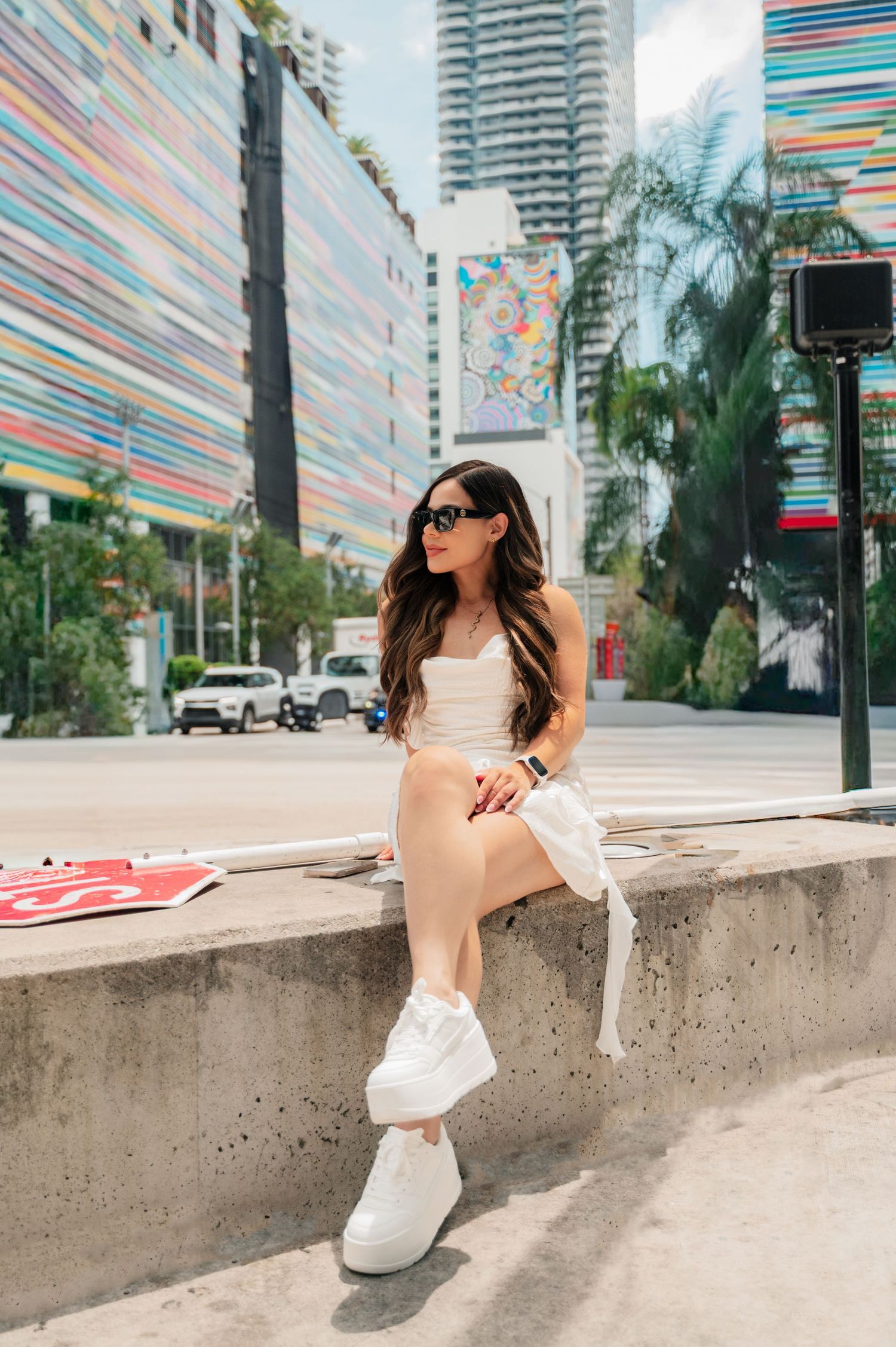 Woman in beige sweater and sunglasses sits on concrete ledge with colorful striped building and palm trees in background.