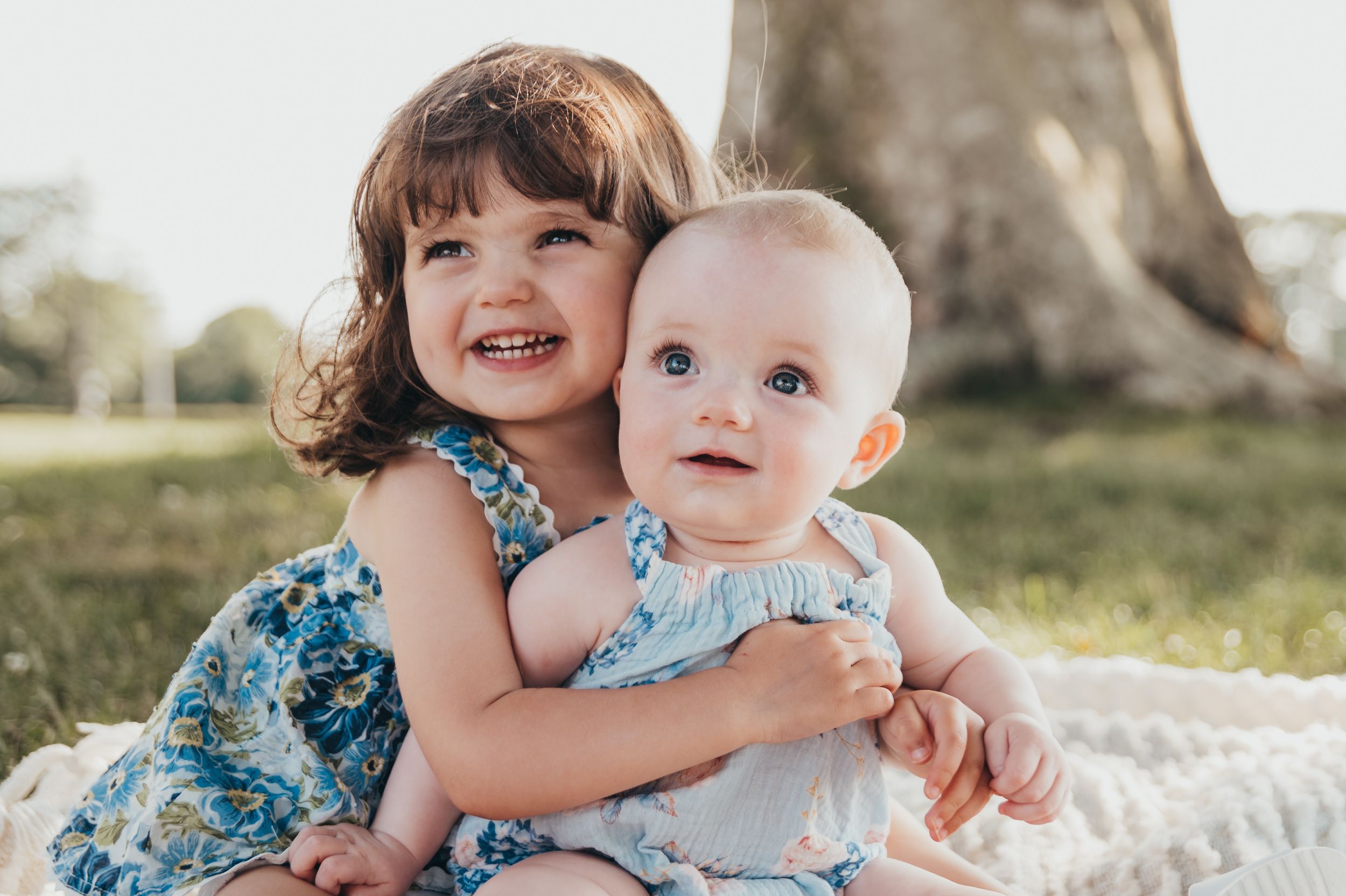 Two children in matching blue floral outfits sit together on a white blanket outdoors in natural lighting.