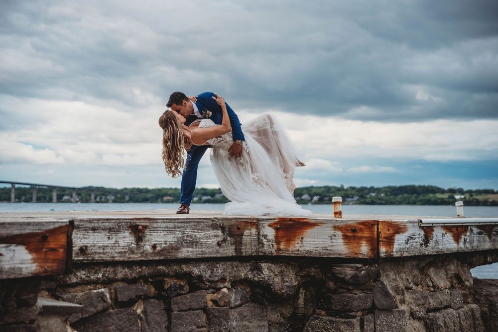 Wedding couple sits on stone wall overlooking scenic waterfront under dramatic cloudy sky.