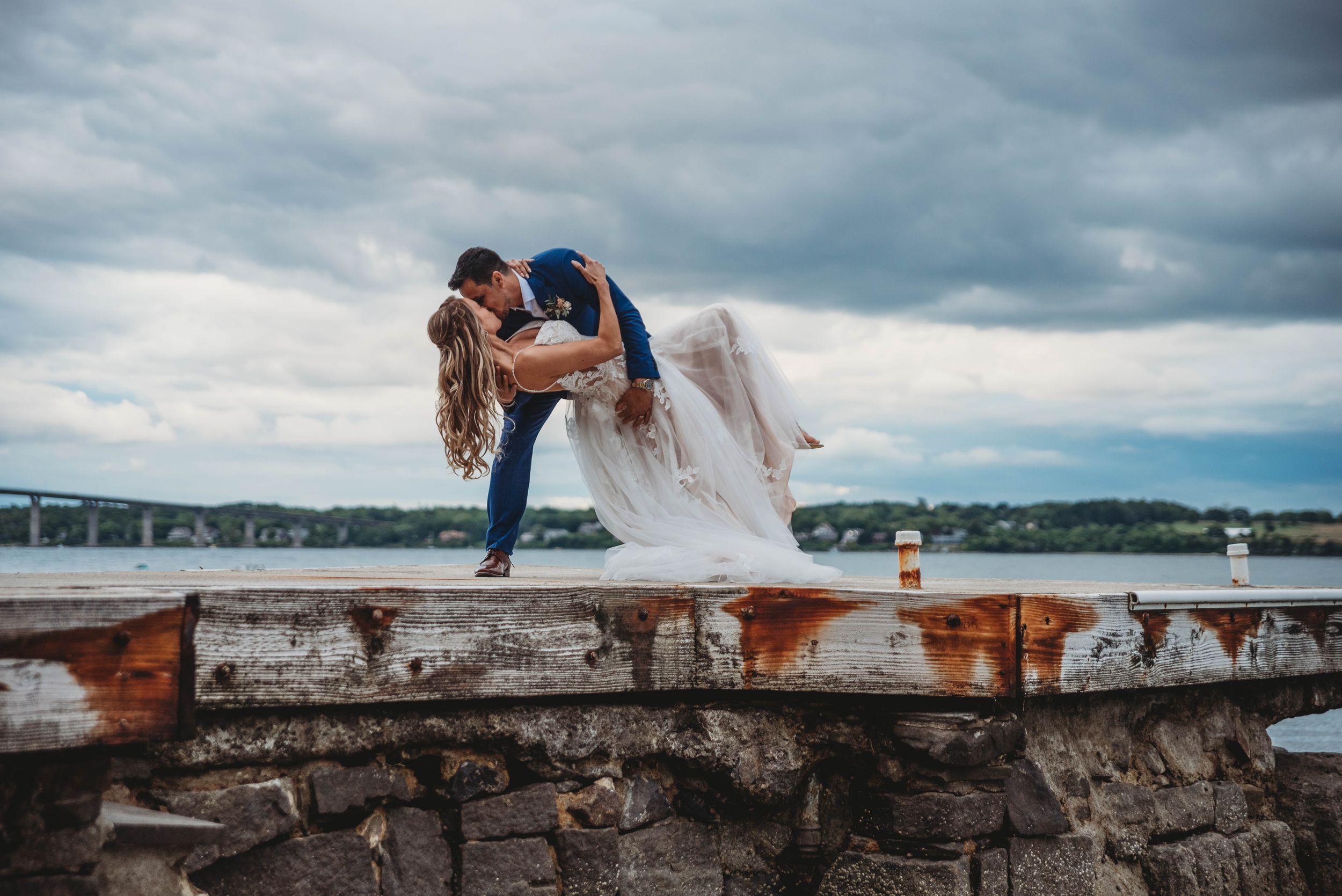 Wedding couple sits on stone wall overlooking scenic waterfront under dramatic cloudy sky.