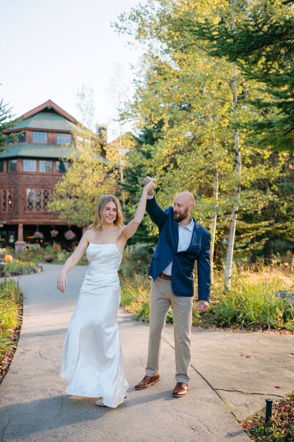 A couple dances playfully on a path with rustic cabin and autumn foliage in the background.