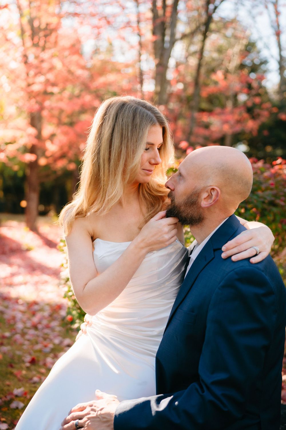 A couple in wedding attire embraces among fall foliage with pink and orange hues.