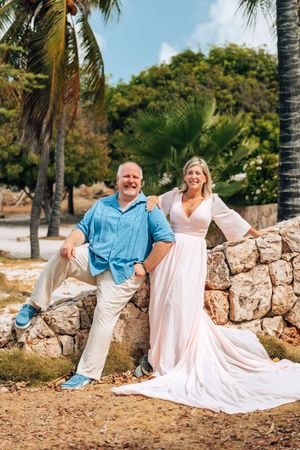 A couple relaxes on a white blanket against a stone wall backdrop with palm trees and greenery in the background.