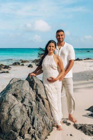 A couple in white outfits poses together on a scenic tropical beach with turquoise water and black volcanic rocks.