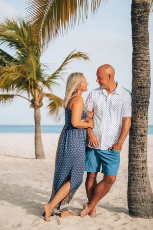 A romantic couple sits together on a tropical beach with palm trees and white sand against a bright blue ocean backdrop.