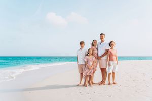 A family in white and beige outfits walks along a pristine beach with turquoise waters in the background.
