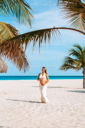 Someone in white clothing walks along a pristine tropical beach with palm trees and turquoise ocean waters.