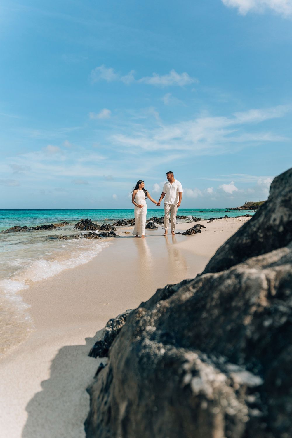 A couple walks along a pristine tropical beach with turquoise waters and black volcanic rocks on a sunny day.