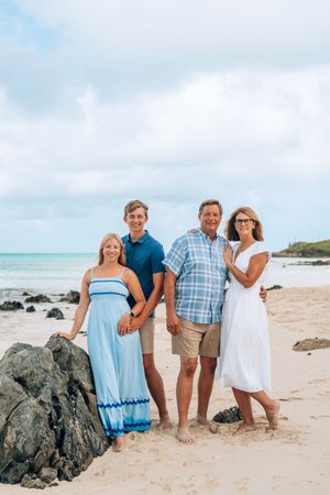 A family poses together on a tropical beach with volcanic rocks and turquoise water in the background.