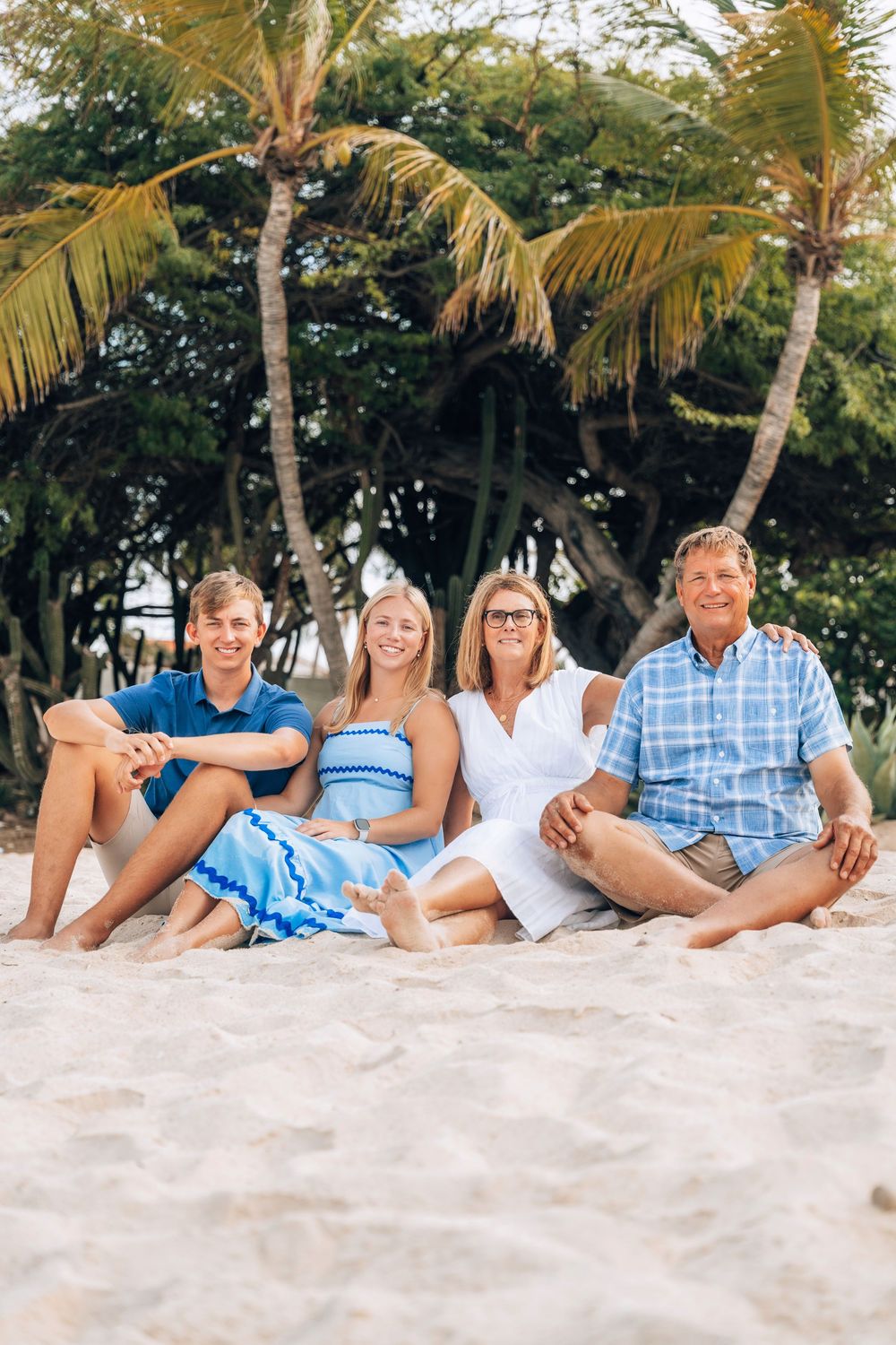 A family group lounges on a tropical beach with palm trees, wearing coordinated blue and white outfits.