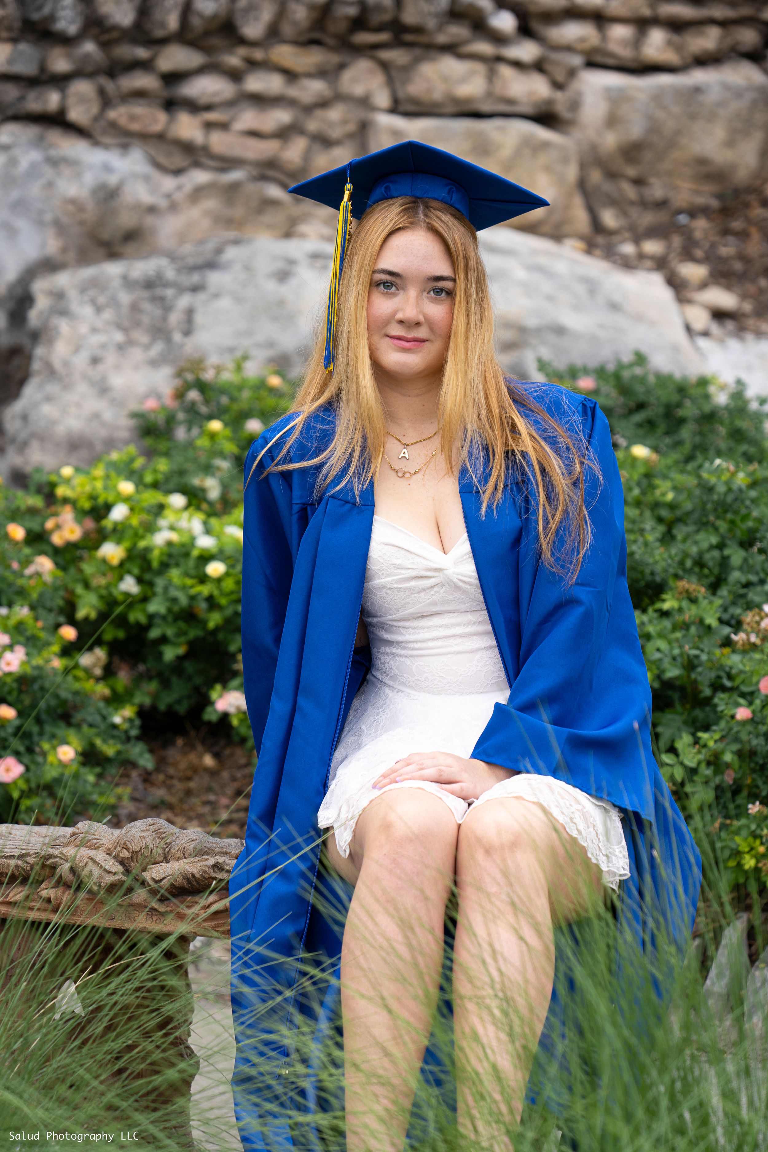 Graduate in blue cap and gown sitting outdoors near stone wall and yellow flowers.