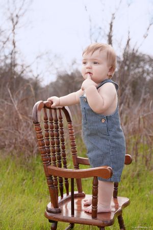 Small child in blue overalls standing on wooden chair in field.