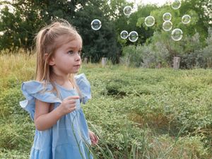 Young girl in a blue dress blowing bubbles in a sunny garden setting.