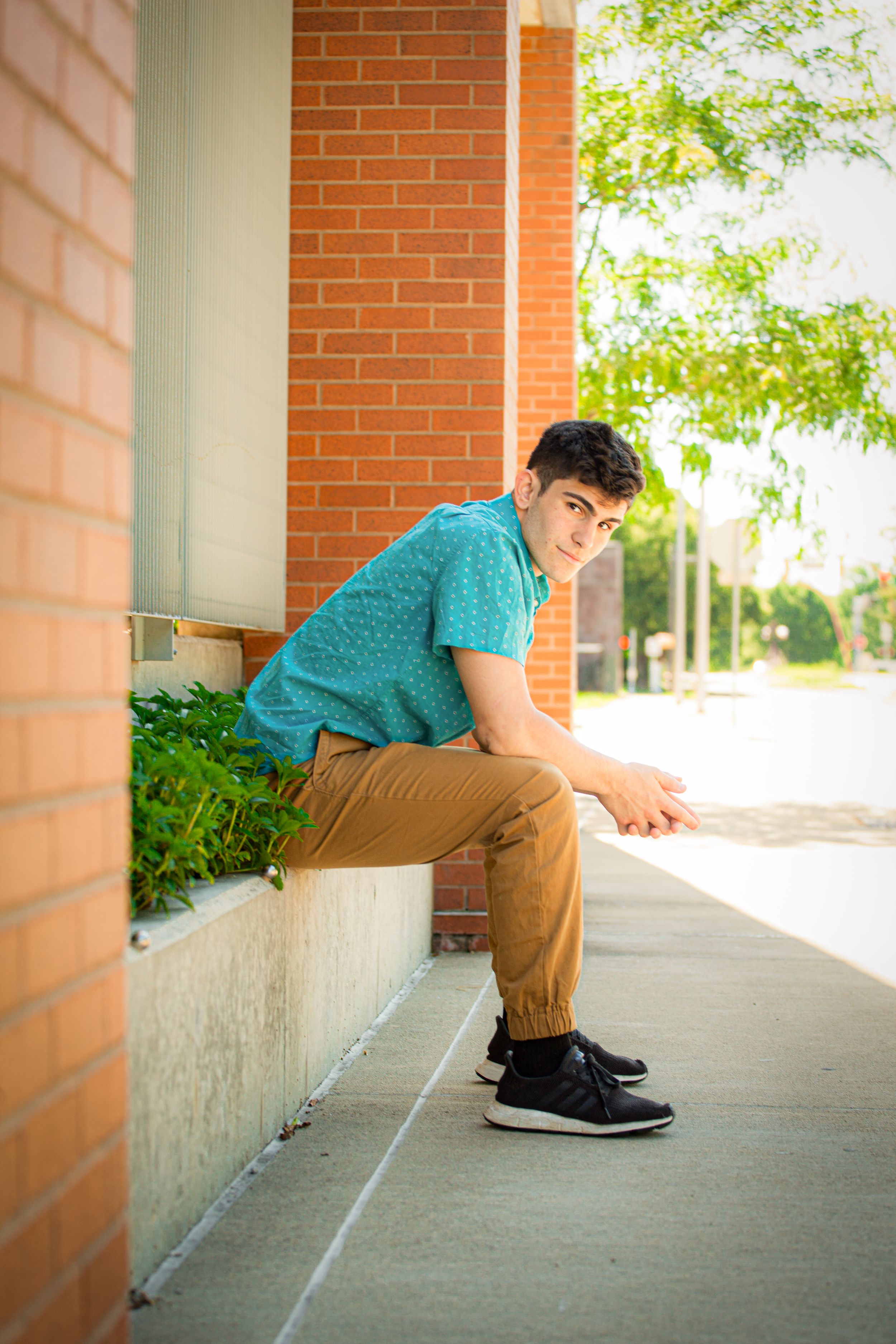 A person in a turquoise polo shirt and khaki pants sits casually on a concrete ledge by a brick building.