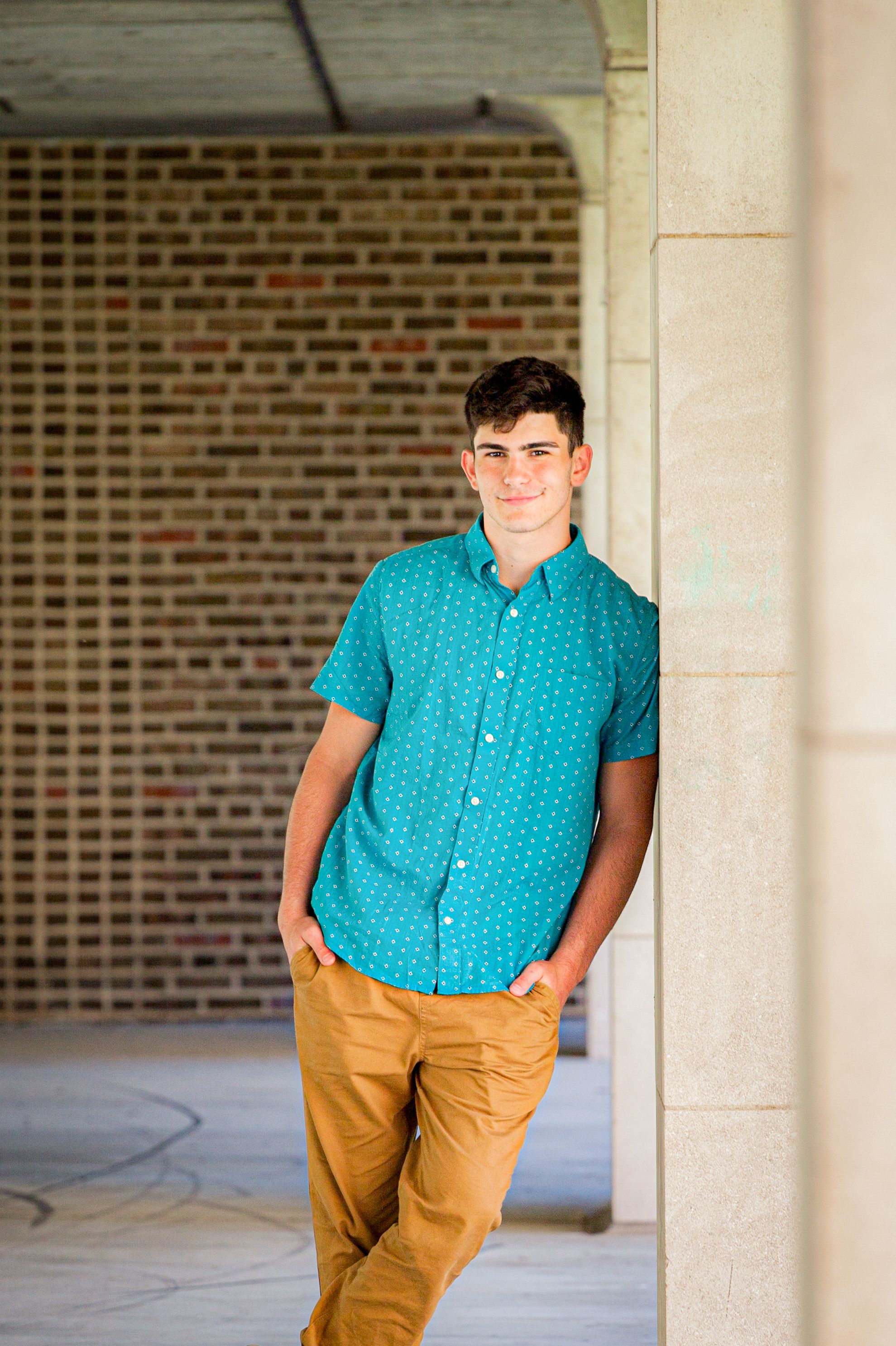 A person in a turquoise button-down shirt and khaki pants poses casually against a brick and stone column exterior.