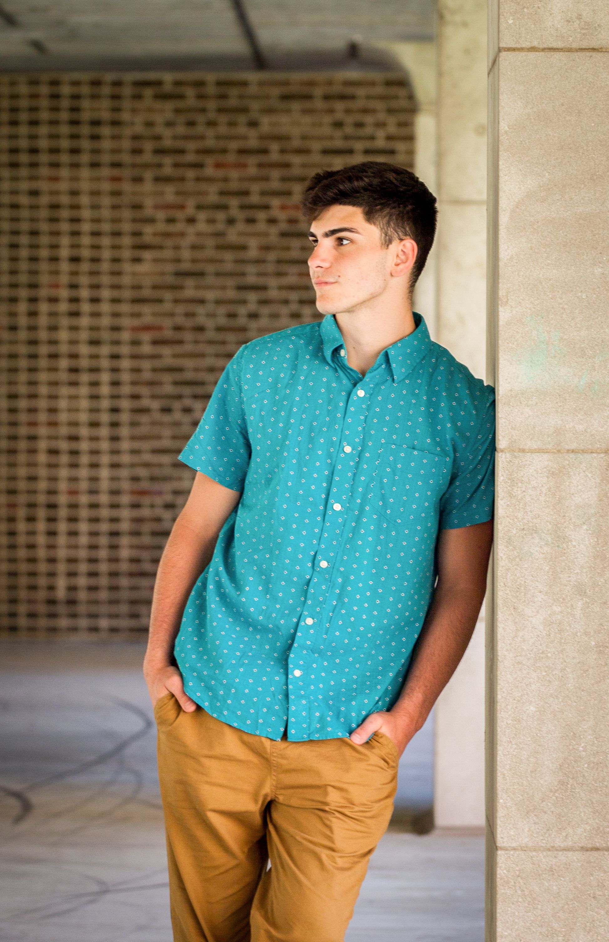 Young person in a turquoise polka dot shirt and khaki pants poses against a stone pillar in an outdoor corridor.