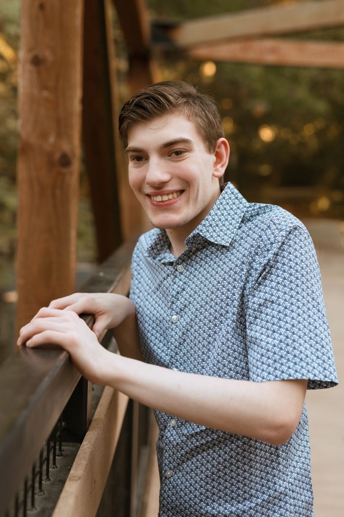 Person in blue patterned short-sleeve shirt leans against wooden post in outdoor setting with warm lighting.