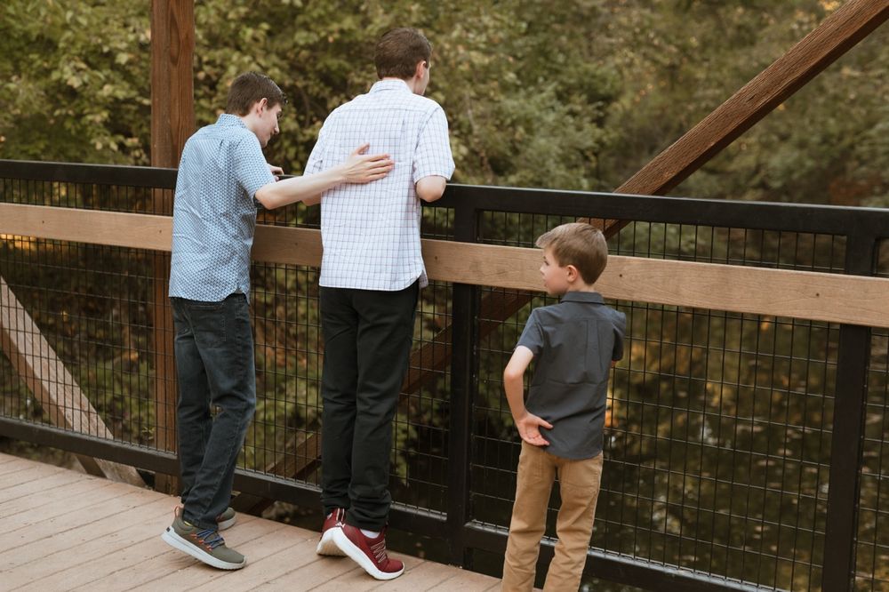 Three people lean over a wooden deck railing to look at something below in a natural outdoor setting.