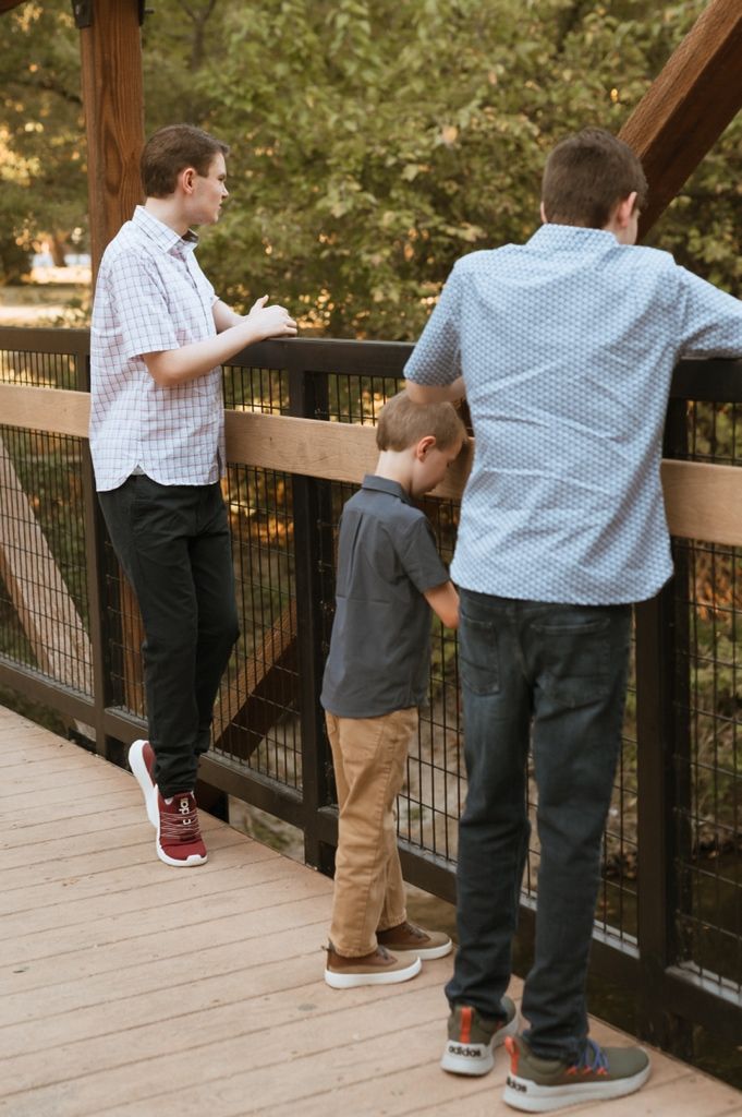Two people stand at a wooden deck railing overlooking trees and greenery on a sunny day.