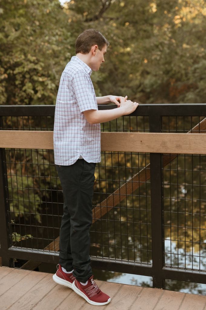 Someone in a checkered shirt and dark pants stands at a wooden deck railing overlooking water at sunset.