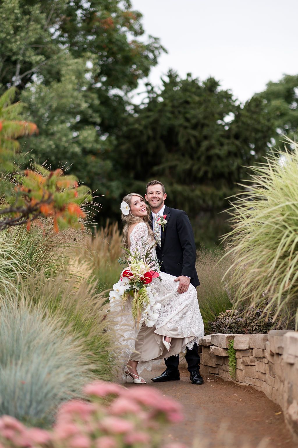 A wedding couple walks through a lush garden path lined with ornamental grasses and flowering plants.