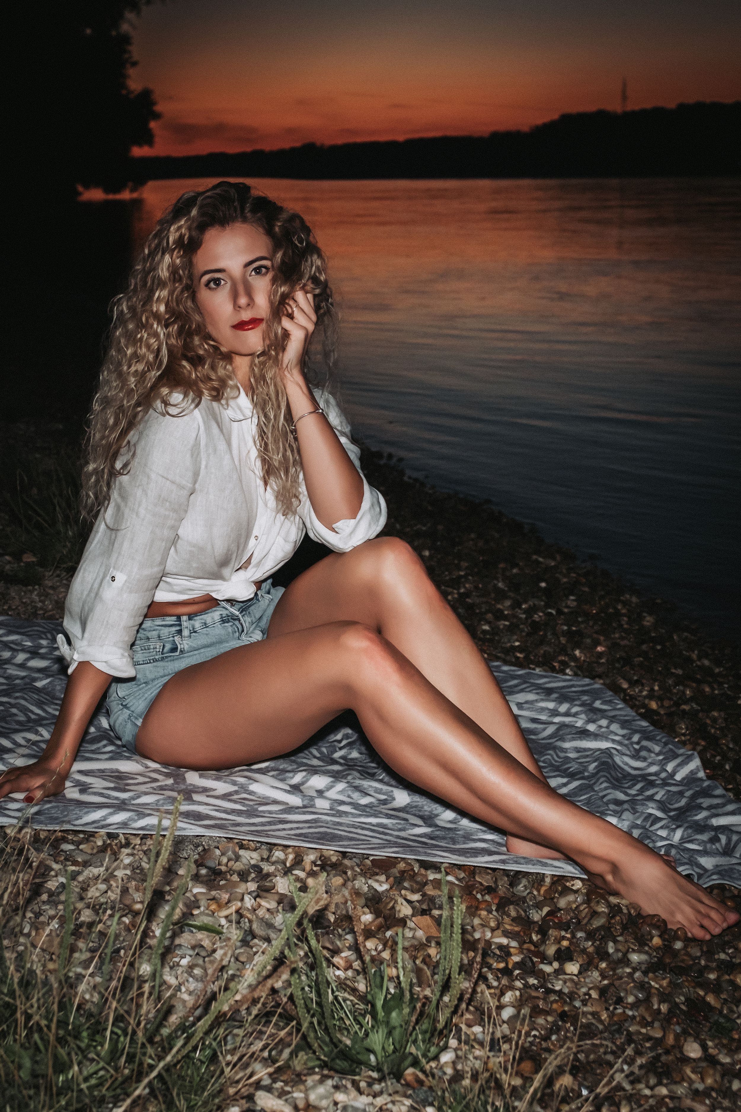 Sunset portrait by the lake with long curly hair and white blouse sitting on blanket during golden hour.