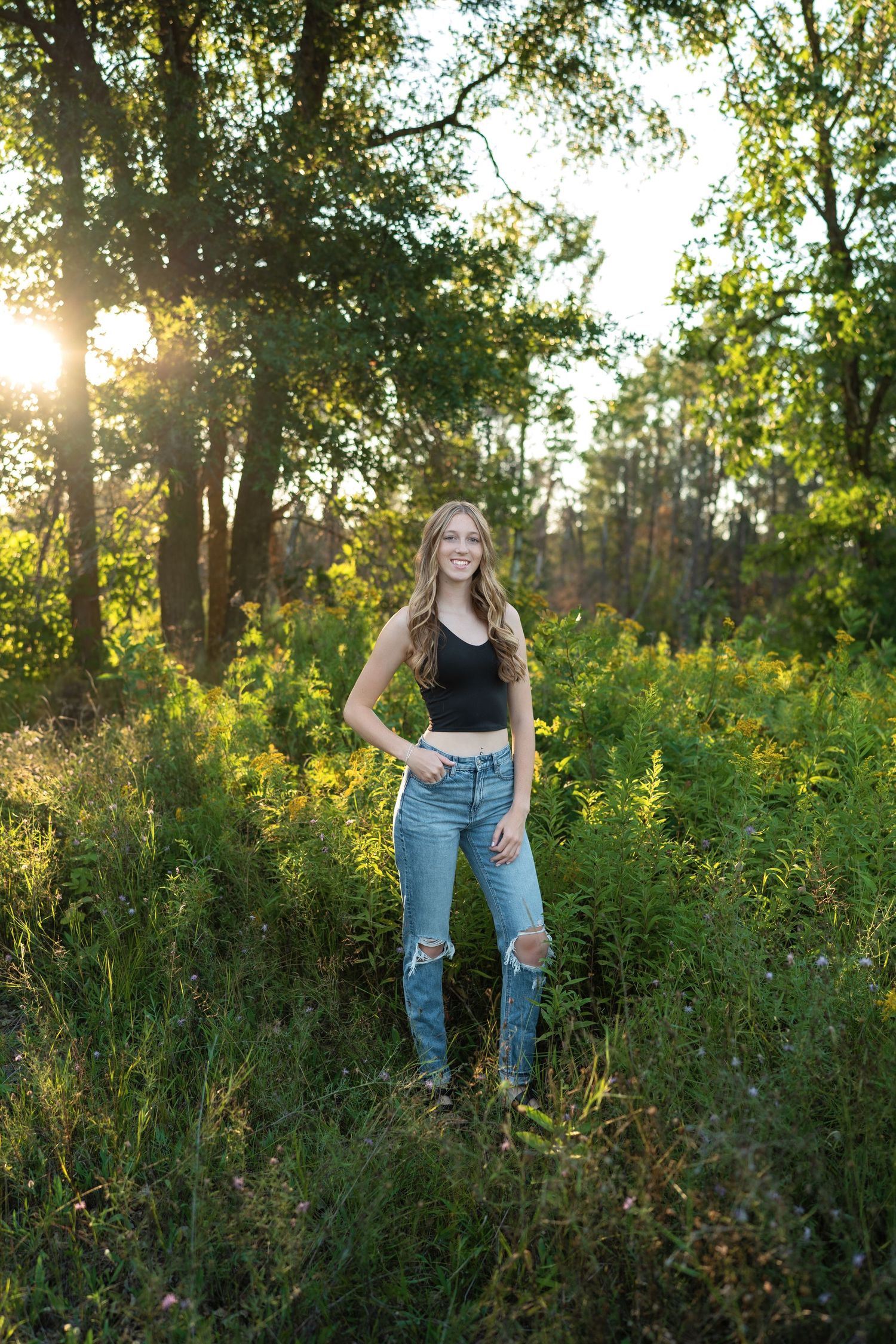 A person stands in a sunlit meadow wearing blue jeans and a black top surrounded by tall grass and trees at sunset.