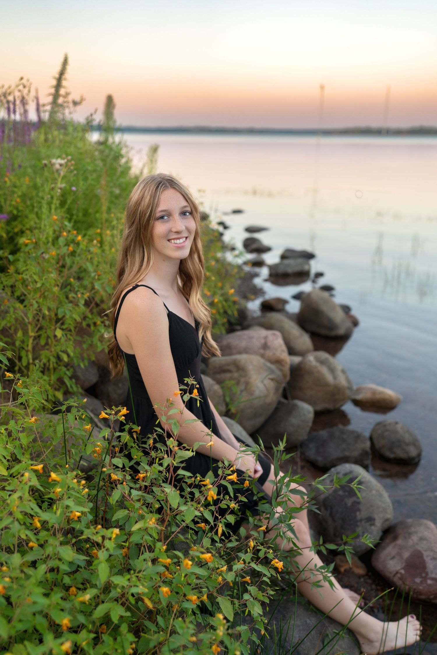 Sunset portrait by a rocky lakeshore with yellow wildflowers in the foreground.