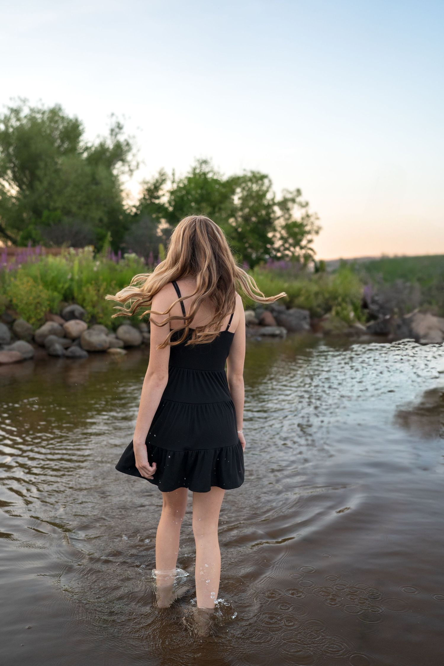 A person wearing a black dress wades through shallow water near a rocky shoreline at sunset.