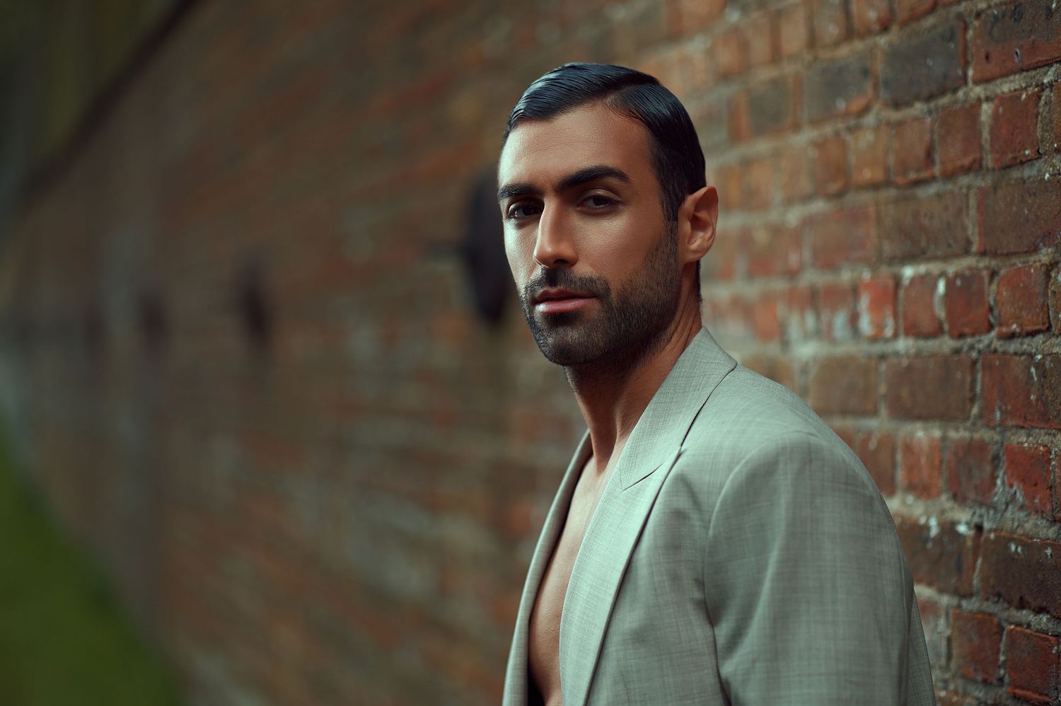 A person in a light gray suit stands against a rustic brick wall in an urban setting.