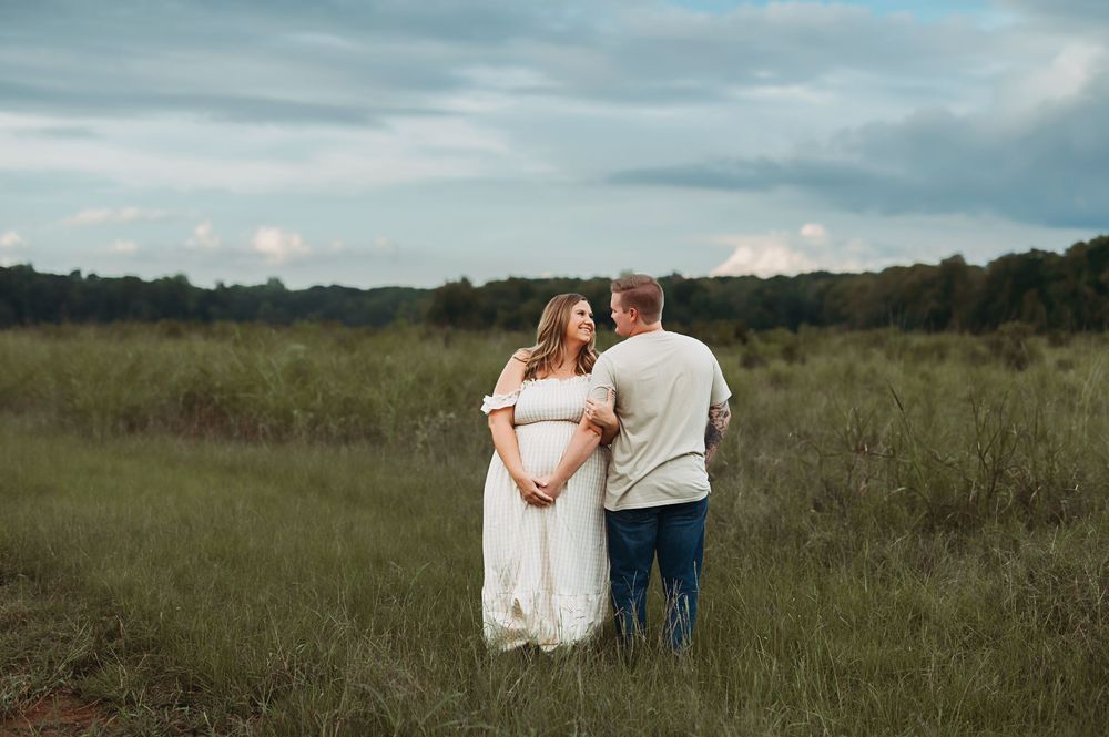 A couple stands together in a serene meadow landscape with cloudy skies overhead.