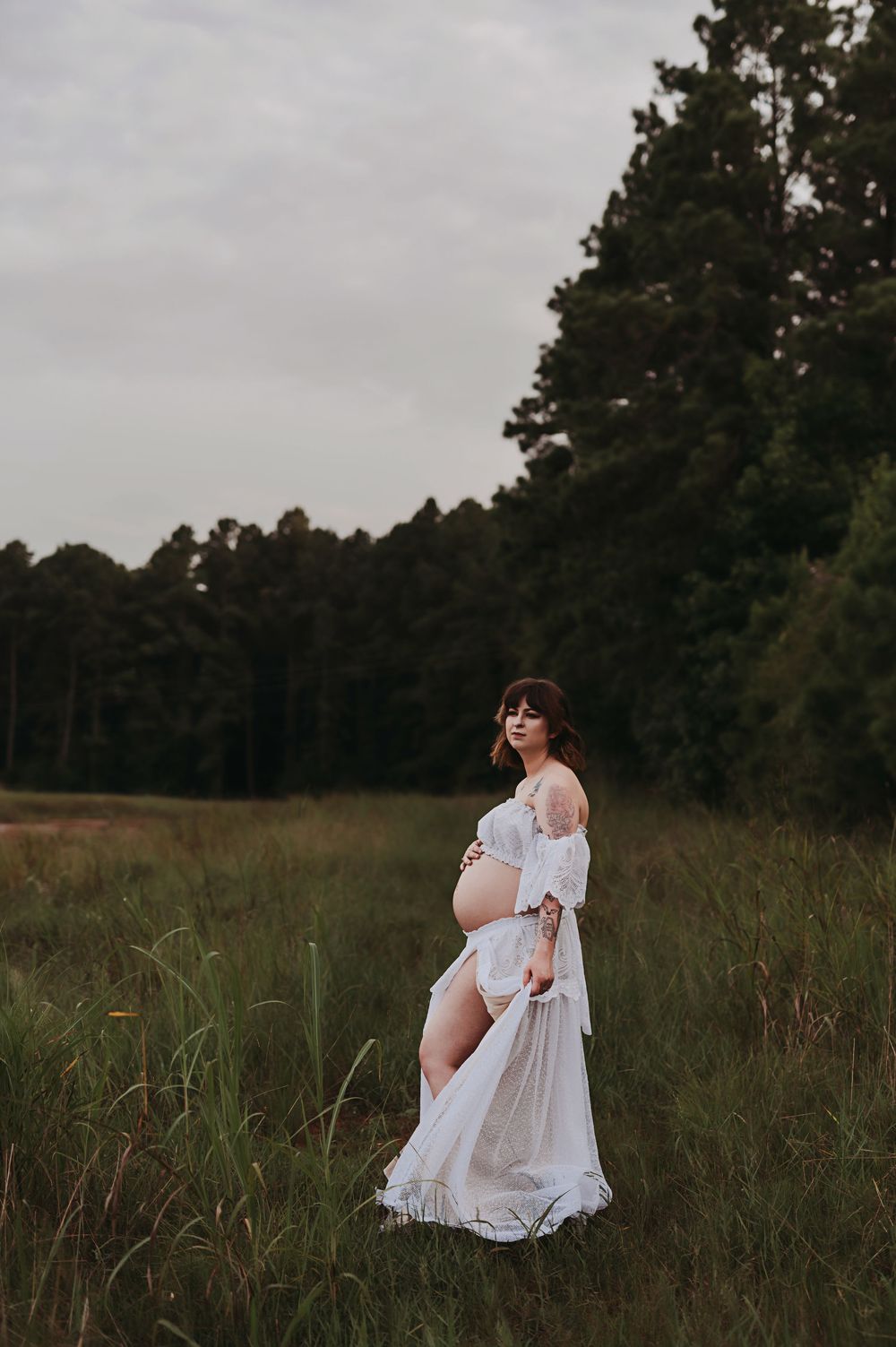 A person in a flowing white dress poses thoughtfully in a serene meadow at dusk with trees in the background.