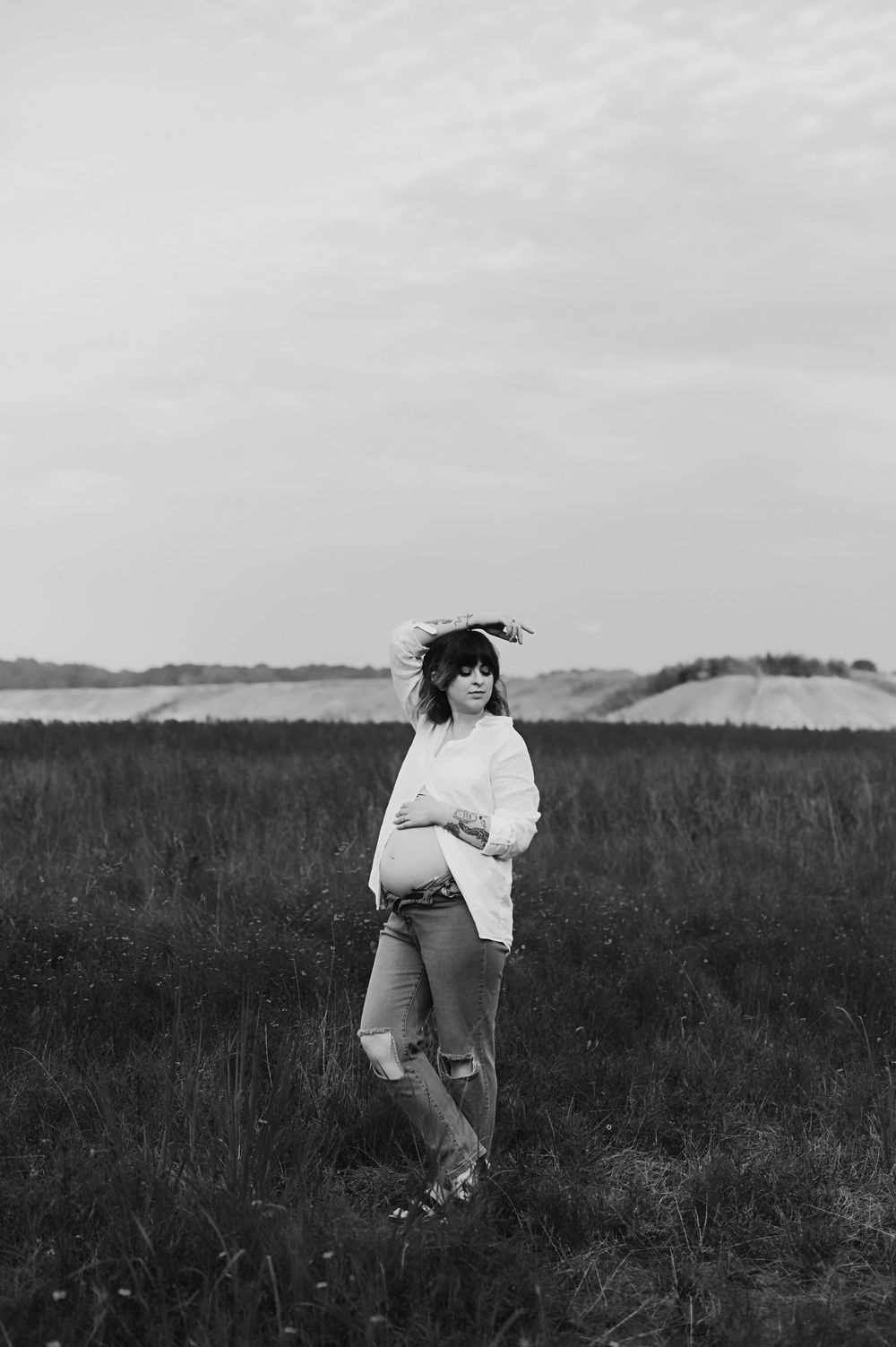 Black and white portrait of someone in ripped jeans and white sweater standing in a grassy field against cloudy sky.