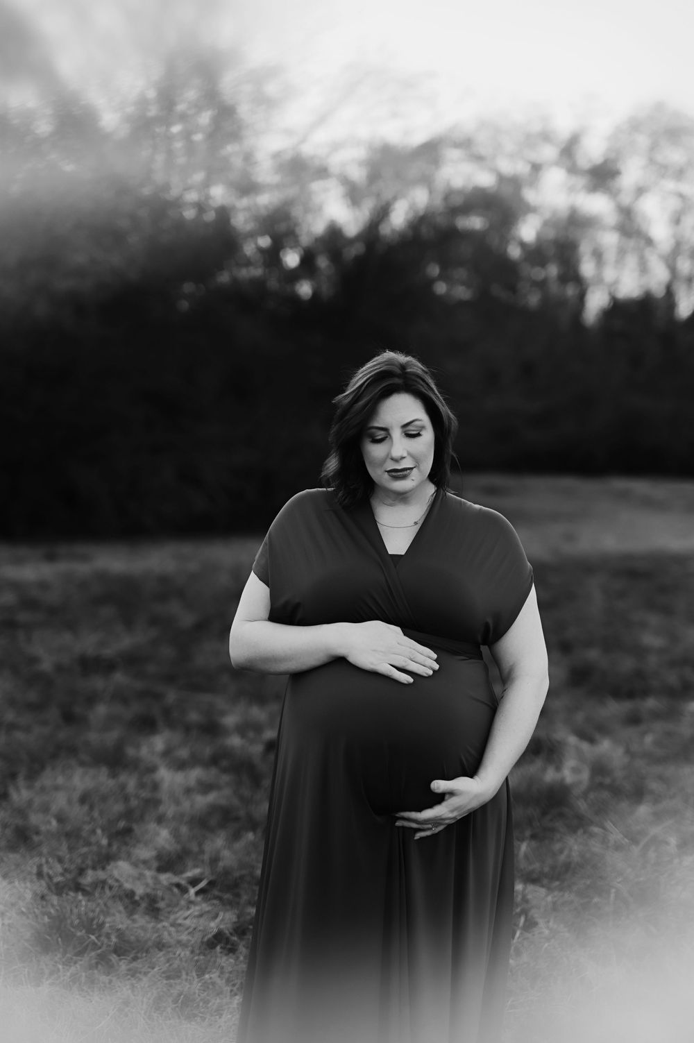 A black and white maternity portrait shows an expectant mother in a dark dress standing outdoors in a dreamy setting in Houston Texas.