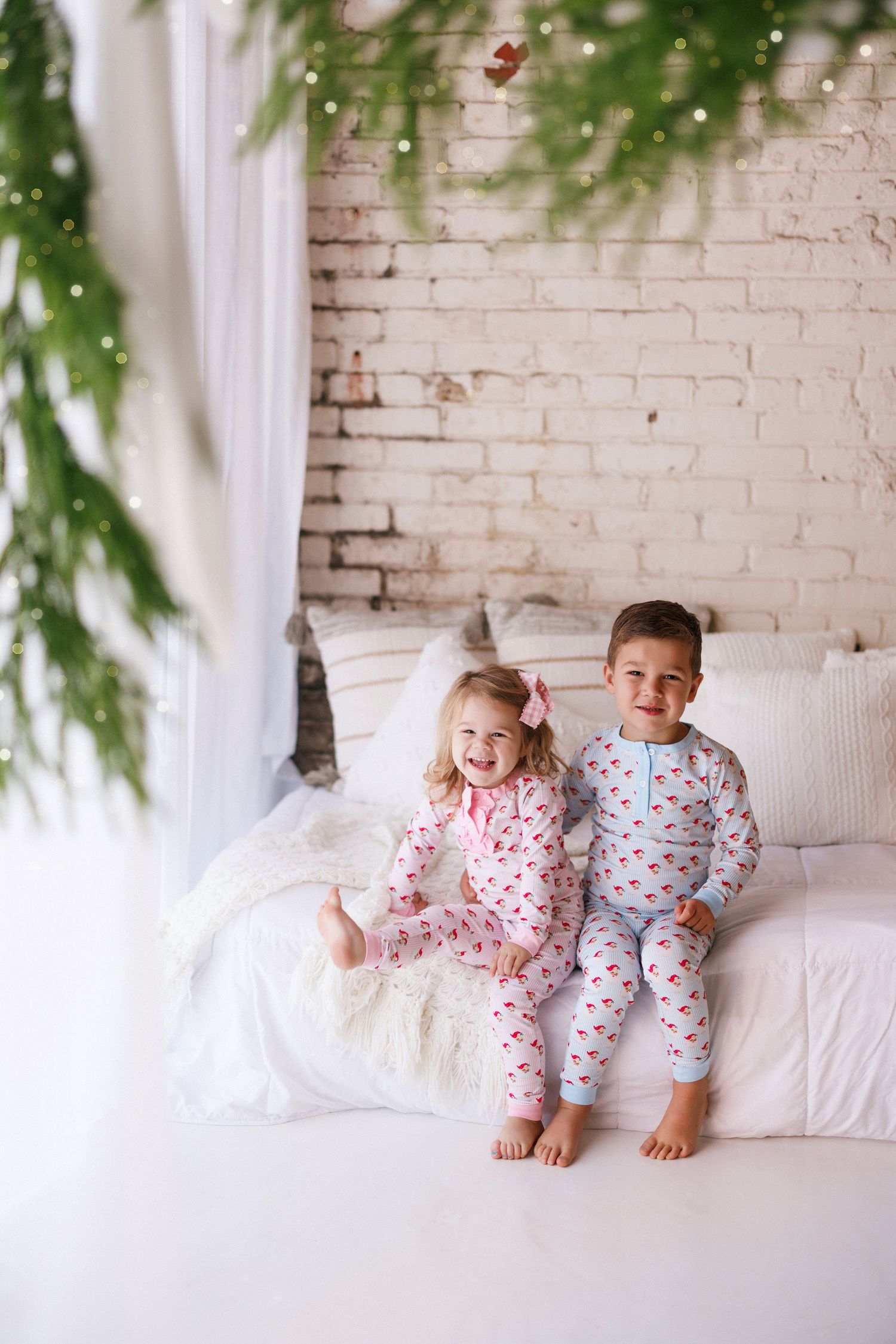 Two children in matching holiday pajamas sitting together on a white couch against a brick wall.