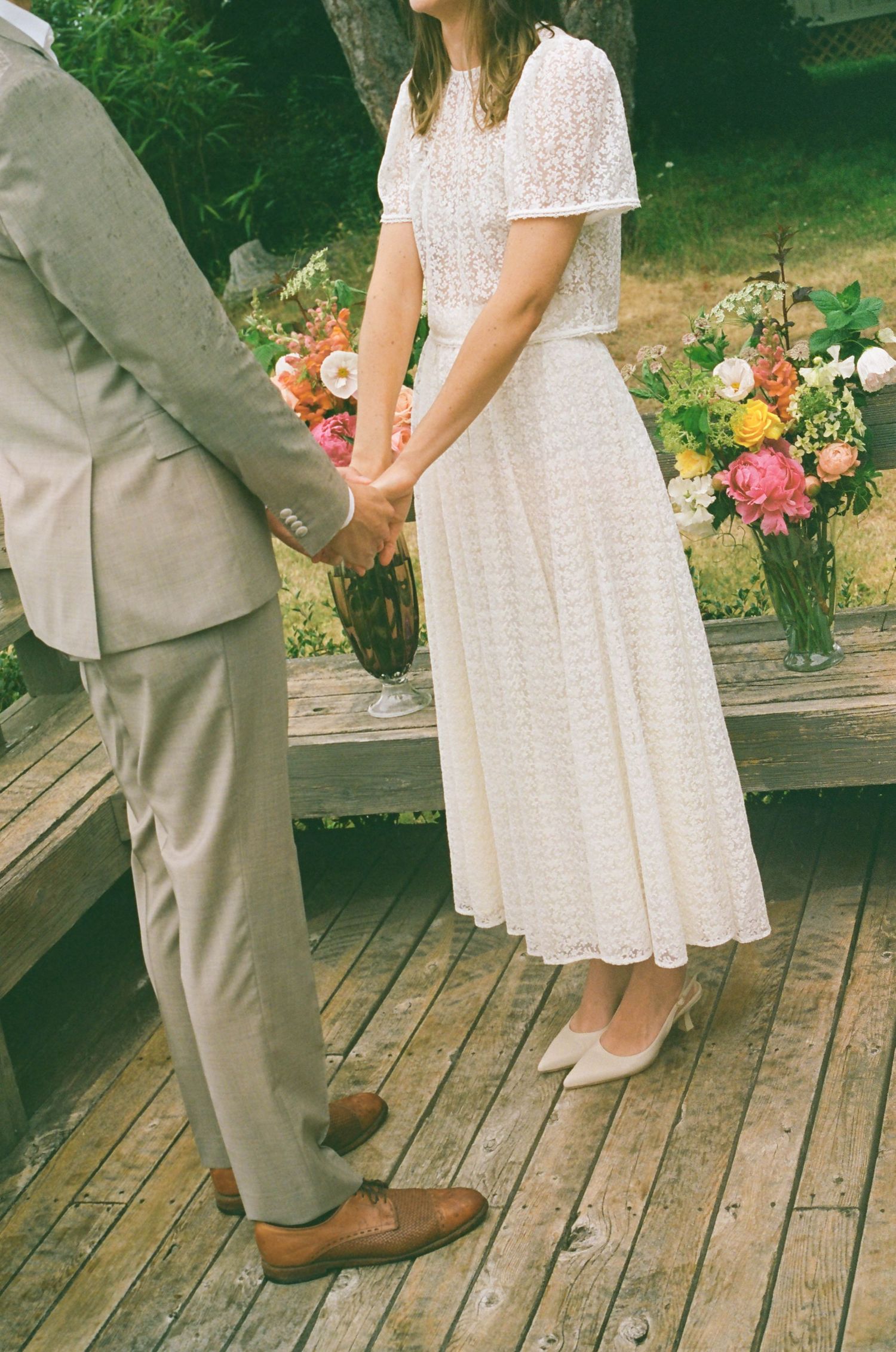 A couple in wedding attire stands on a rustic wooden deck with colorful flowers in the background.
