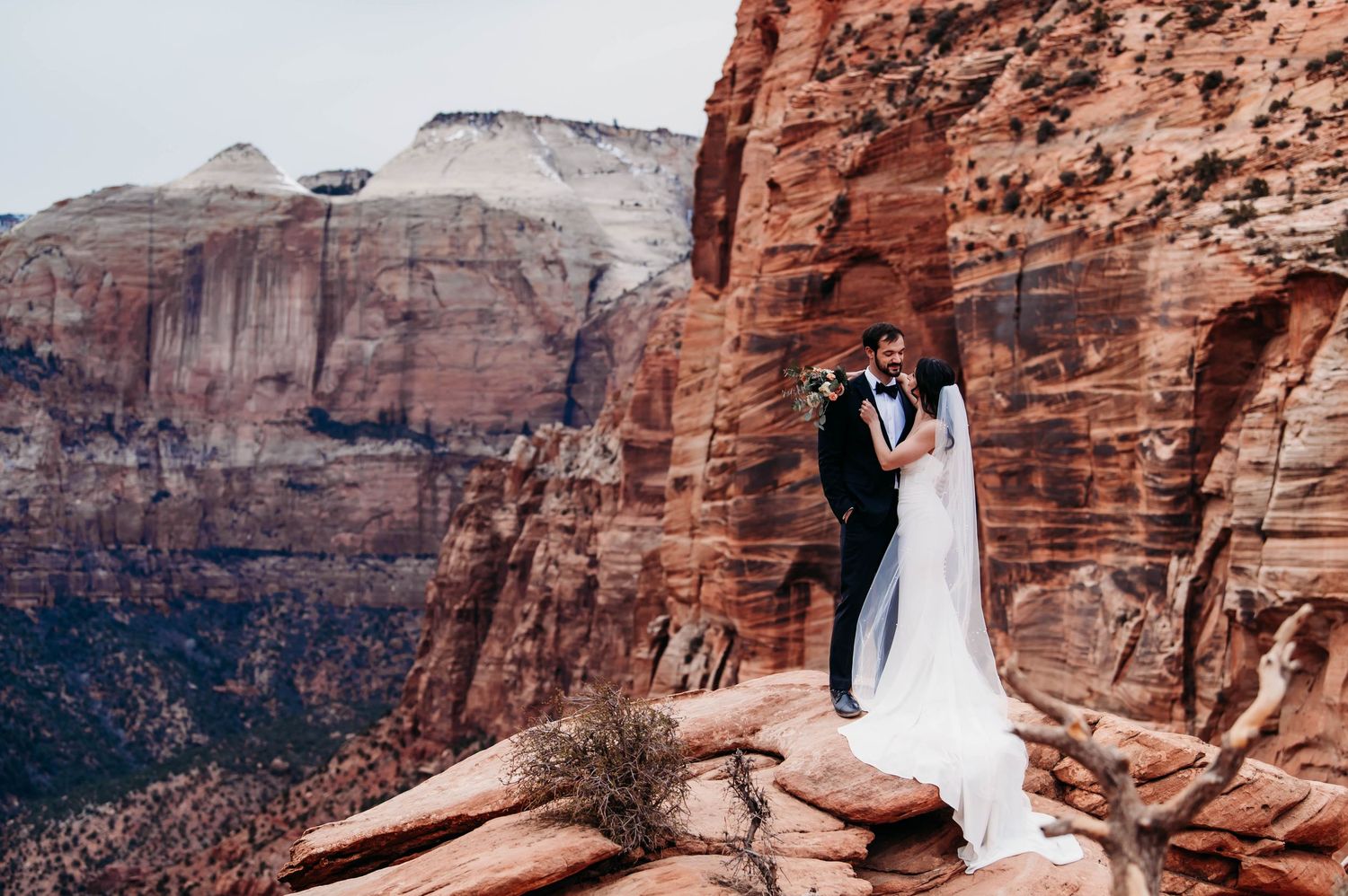 A romantic couple stands together in a golden autumn field with dramatic cloudy skies and mountains in the background.