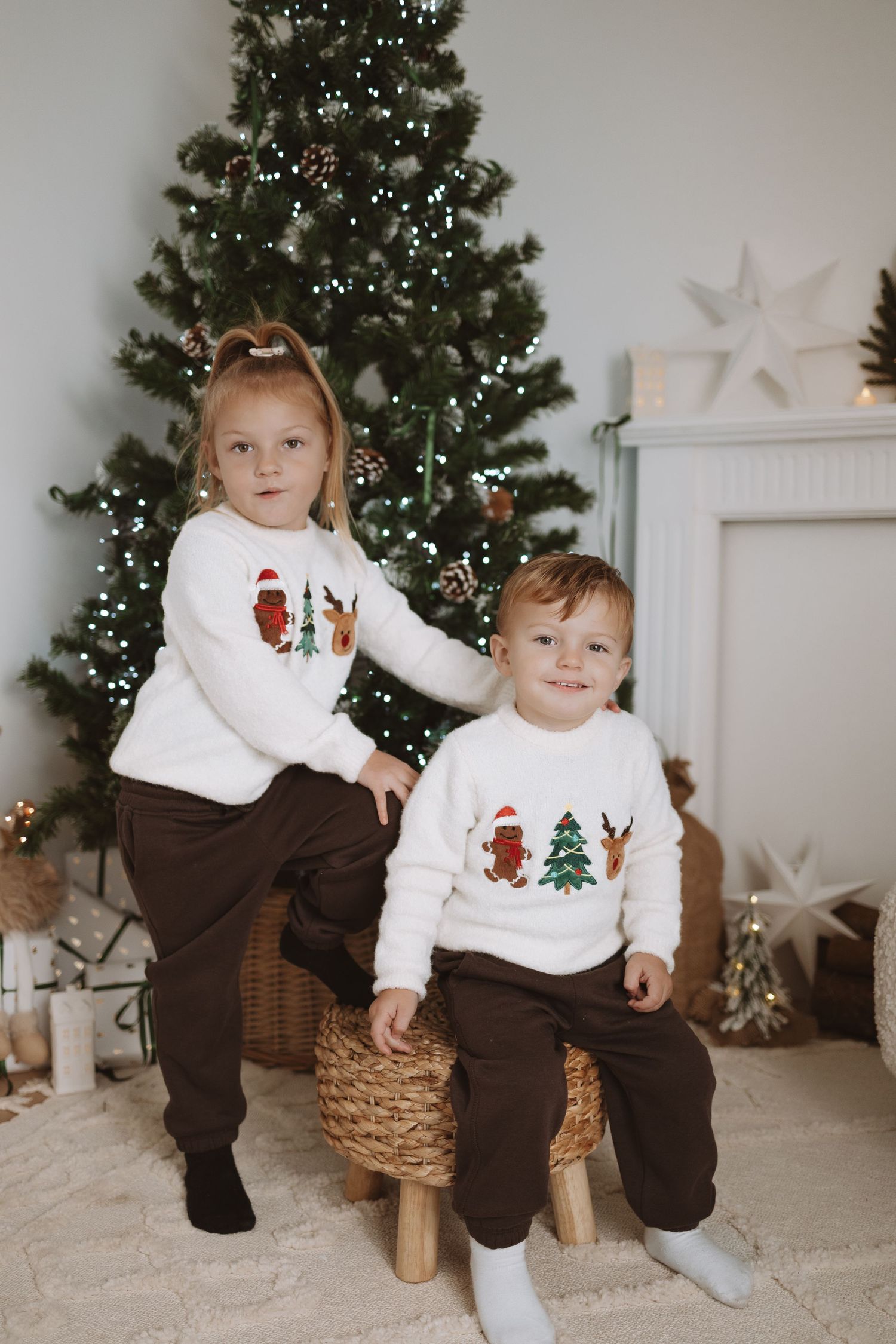 Children in matching white holiday sweaters and brown pants pose in front of a decorated Christmas tree.