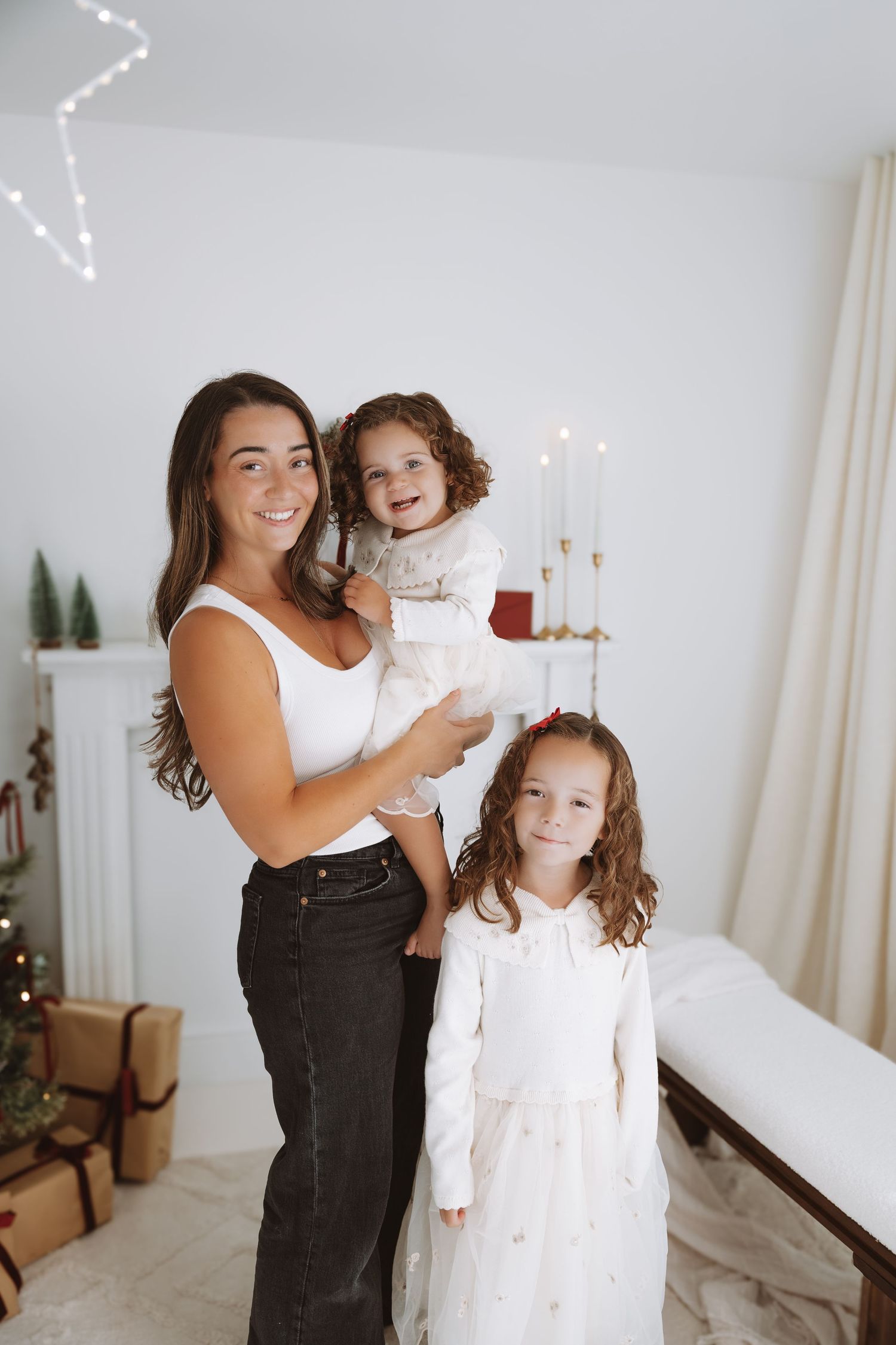 Children in matching white holiday sweaters and brown pants pose in front of a decorated Christmas tree.