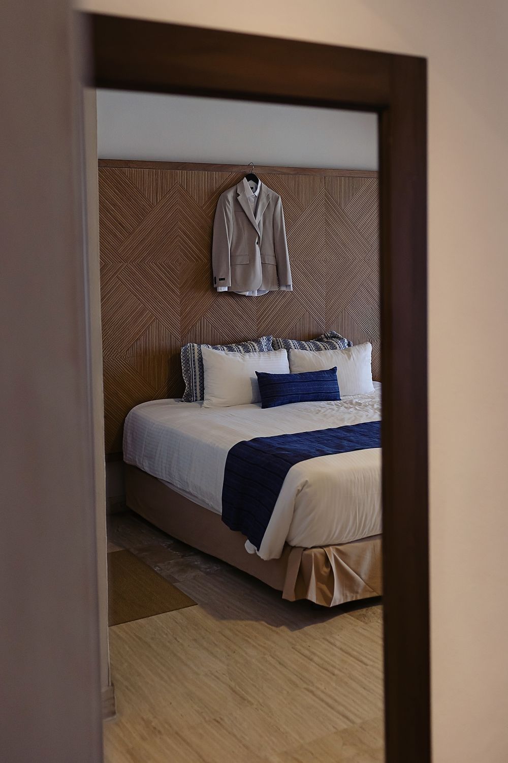 Modern hotel room interior with white bedding, navy blue accents, and a gray blazer hanging on textured wallpaper.