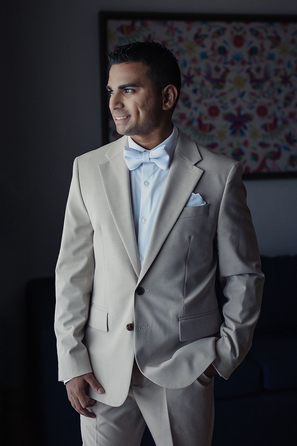 A groom wearing a light gray suit with a light blue tie and pocket square poses against a dark background.