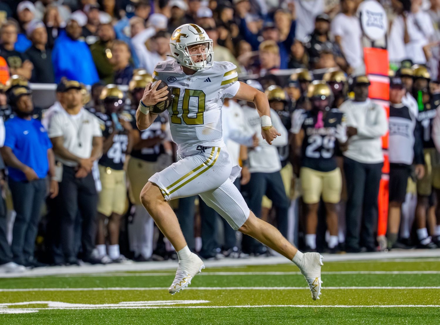 Football player in white jersey dives forward to make a catch during a game with spectators in background.