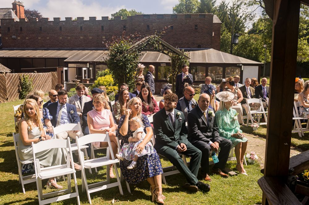 Wedding guests seated in white chairs during an outdoor ceremony under trees.