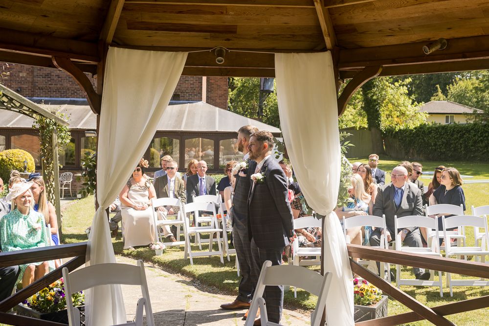 Wedding ceremony under a white draped gazebo with guests seated on white chairs.